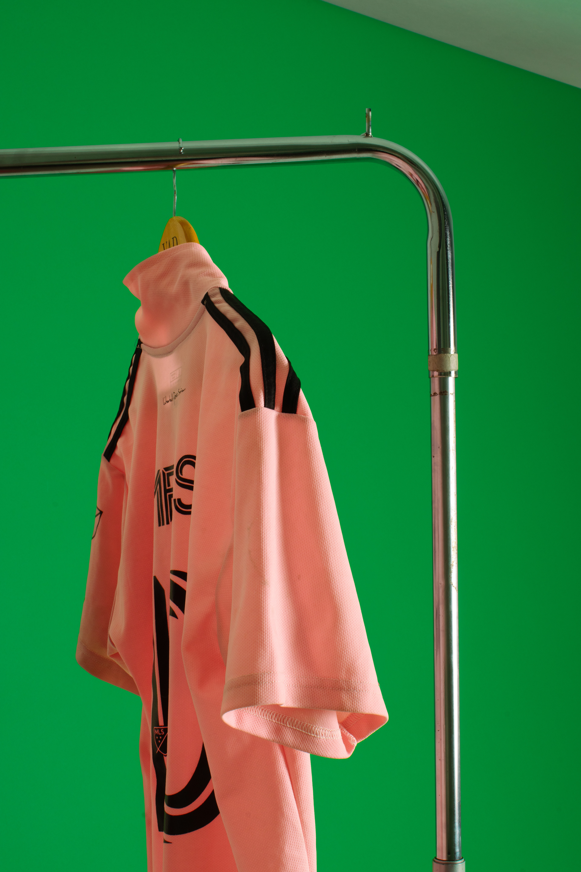 Camiseta rosa de fútbol del Inter Miami colgada de un percha amarilla en un caño de perchero cromado, sobre fondo de croma verde.