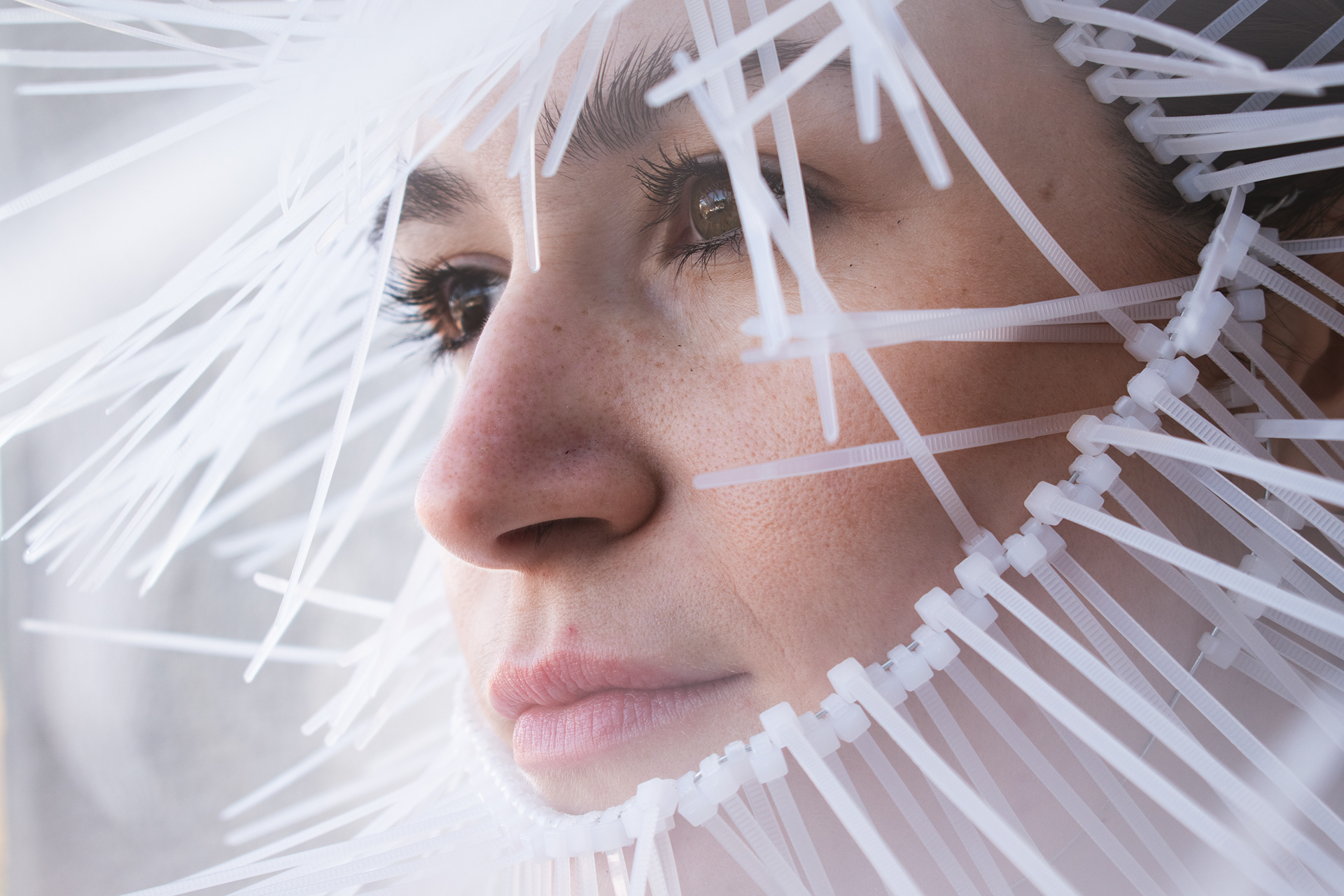 Primer plano del rostro de una mujer de perfil rodeado por un collar-artefacto de precintos plásticos blancos, cuyas puntas se proyectan hacia afuera como púas defensivas.