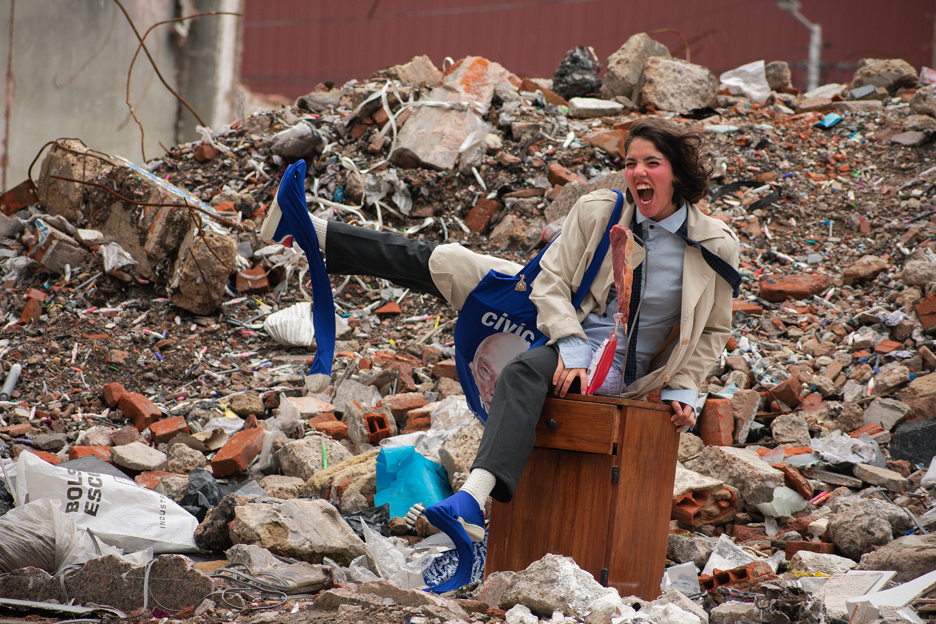 Mujer con impermeable beige y camisa con corbata grita sentada sobre una mesa de luz de madera en medio de un descampado de escombros, con un bolso azul de campaña política cruzado y un trozo de carne cruda colgando del pecho.